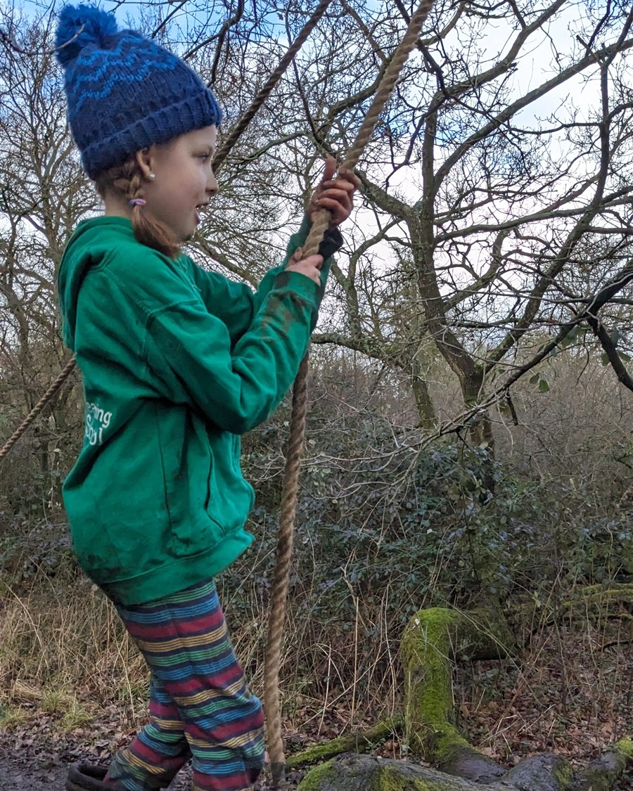 A child in a green jumper and bobble hat confidently using a rope swing in the winter woodland