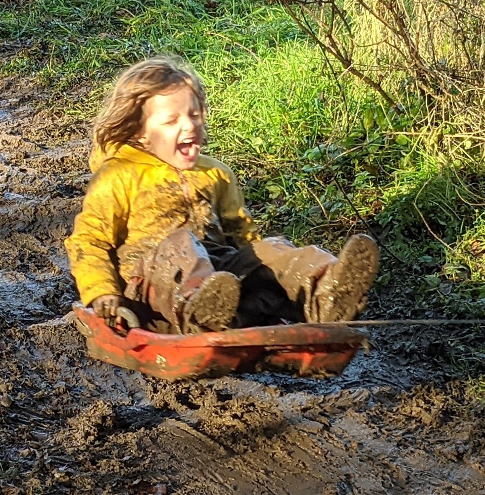 A delighted child in a yellow waterproof coat sledging through the mud
