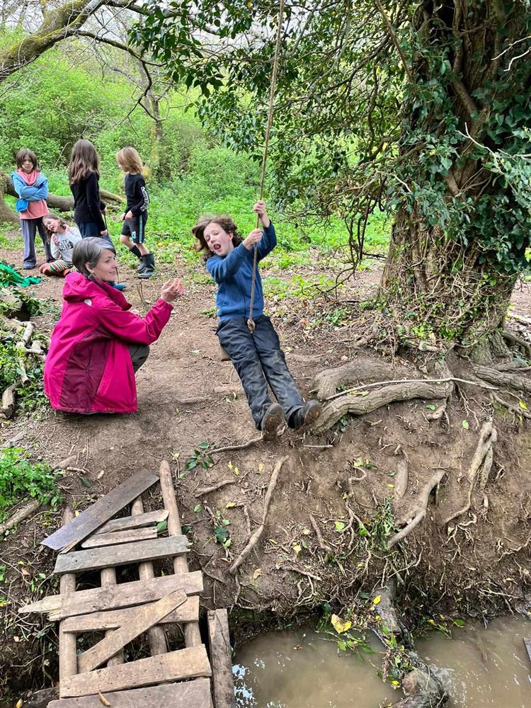 A child swinging across the stream on a rope swing while a leader cheers them on