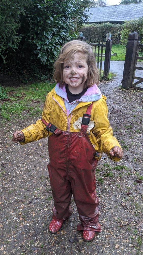 A happy child with a muddy face in a yellow waterproof coat and red waterproof dungarees
